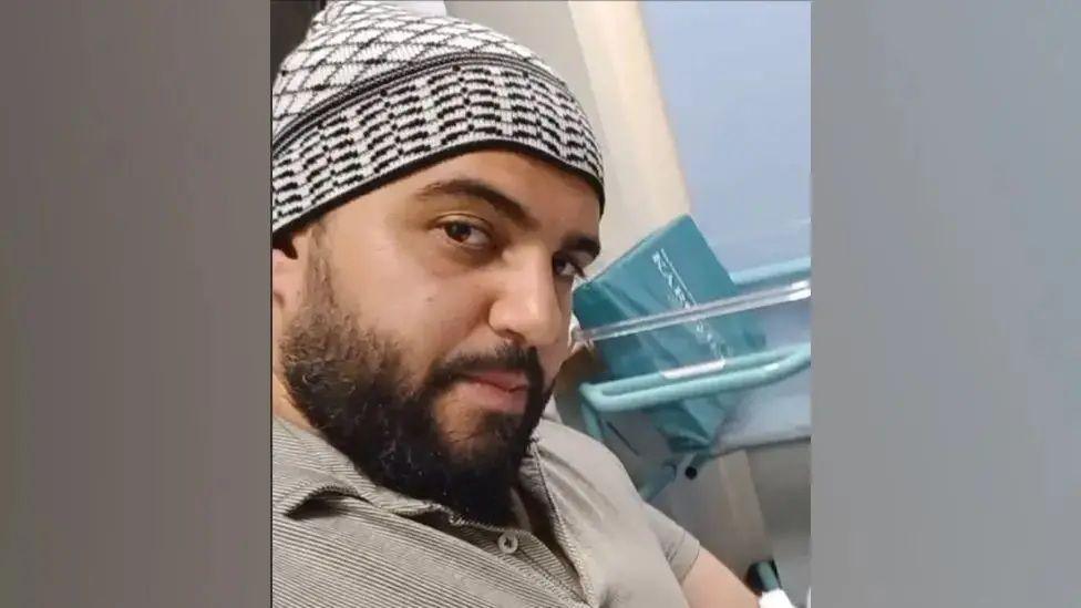 A selfie of a man who is sitting down in a hospital. He is looking directly at the camera. He is wearing a beige polo shirt and wearing a black and is wearing what appears to be a white knitted kufi hat.