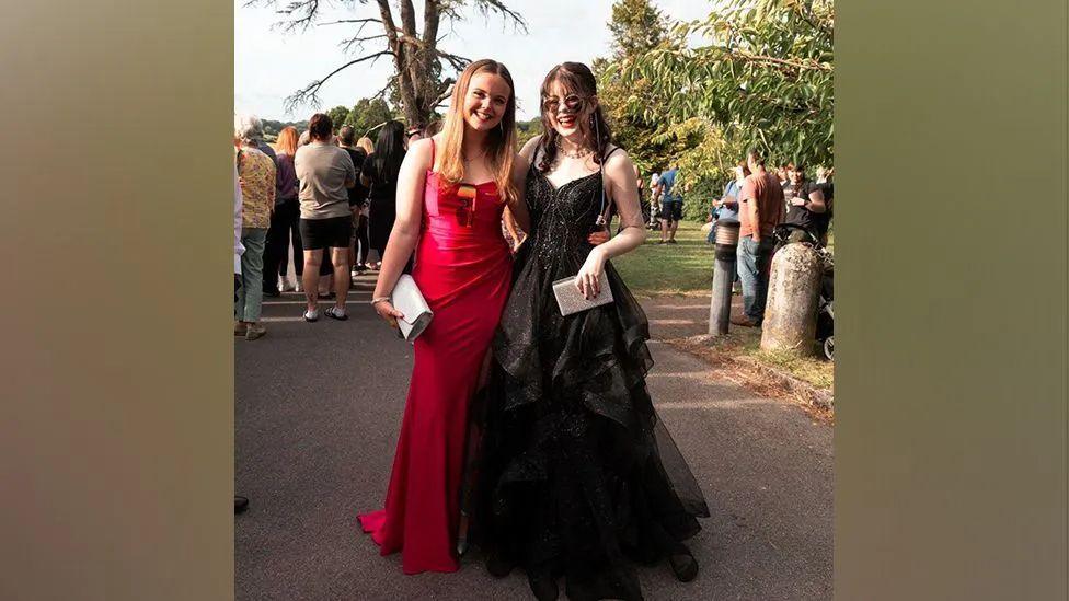 Ellie in a long black prom dress with a friend in a red prom dress and both carrying clutch handbags smiling for the camera