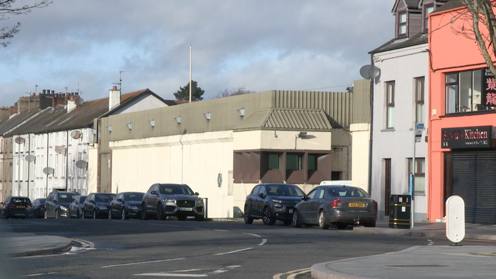 A white police station with brown windows. There are several black and cars outside the station on a road. Other white buildings are visible beside station, a grey and orange building are on the other side