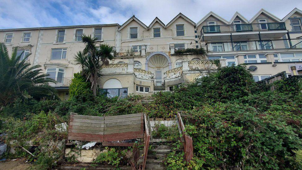 Large cream hotel building fills the picture with collapsed wooden steps in front and sprawling vegetation.