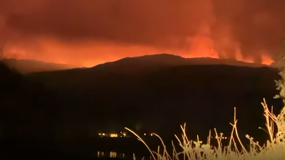 A dark hill at night with a glowing orange red sky showing a fire in the distance.