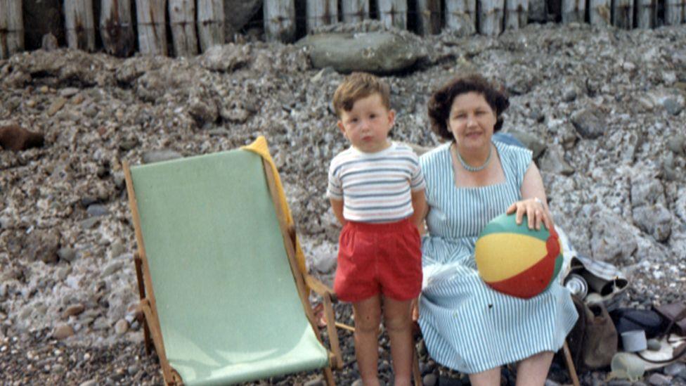 A very young Dave Myers is standing next to his mother on the beach. He is wearing a striped t-shirt and red shorts. His mother is sitting in a deckchair next to him and she is wearing a stripy blue dress holding a beach ball. She has brown hair. 