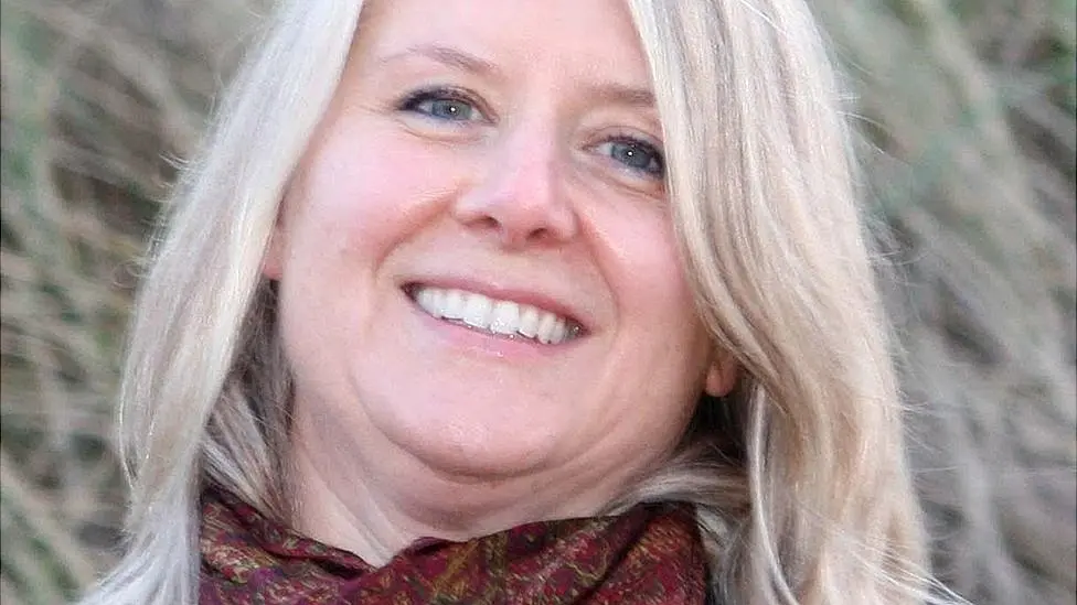 Joanna Simpson has shoulder length blonde hair and is wearing a red patterned scarf. She is smiling at the camera 