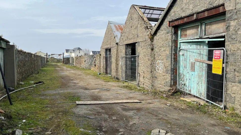 There are three empty warehouses on the right, with metal fencing across the entry ways. There is a scruffy, untarmacked road in front of them. 