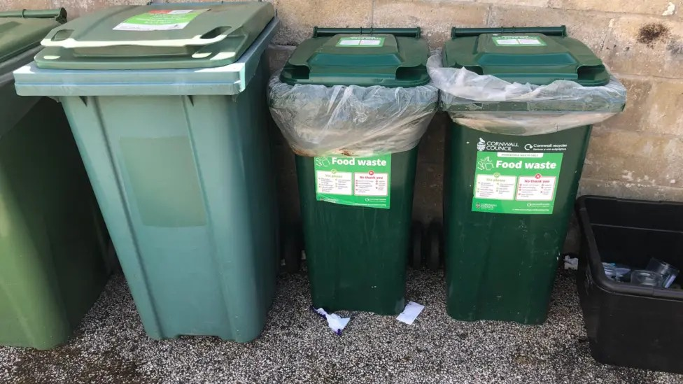 Several green rubbish bins, food waste bins and a black recycling box filled with glass in front of a breeze block wall.