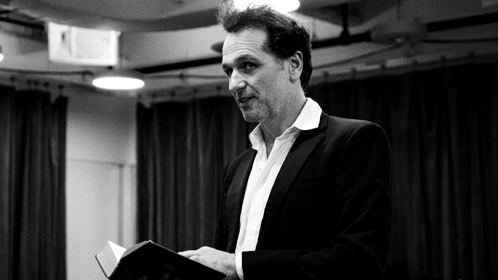 Black and white image of Matthew Rhys in rehearsals as Burton. He is wearing a white shirt, black suit and is holding a book. He is facing forward, away from the camera.