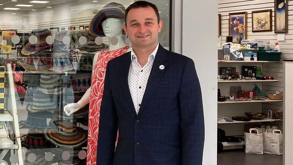 A man, Dawid Kozlowski, has short brown hair and is wearing a dark blue jacket and white dotted shirt. He is standing outstanding a shop with hats, clothes and bric-a-brac inside.