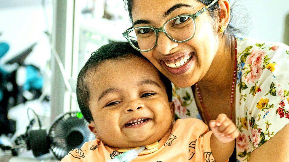 Little Gabriel is smiling and looking at someone off camera. He is wearing a beige top and you can see a breathing tube in a hole in his neck. His mum is also smiling broadly and looking at the camera while holding her son's head up. She is wearing a floral top and green glasses.