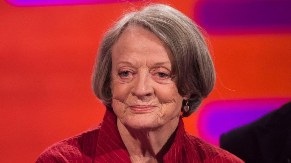 Dame Maggie Smith, a woman with dark grey bobbed hair, wearing a red shirt against a red and purple backdrop 