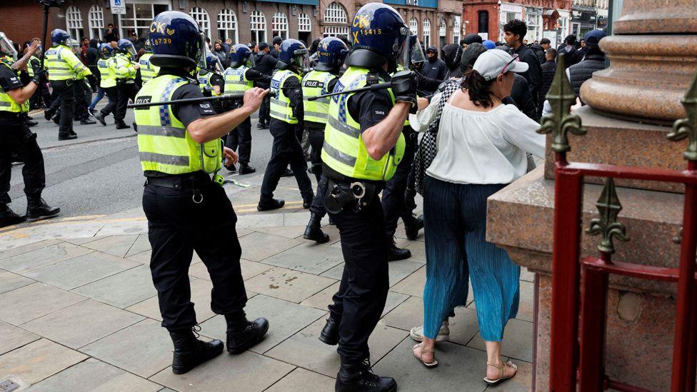 Two police officers in riot gear and holding batons on their shoulders, stand close to a woman in a white cap, white top and blue trousers as a line of police officers pushes a group of angry-looking men back down a street