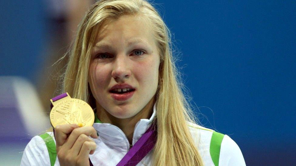 Headshot of Rūta Meilutytė at her medal ceremony in 2012. She has long,straight hair and is wearing a white, zipped tracksuit top. She is holding up a gold medal with a purple ribbon.