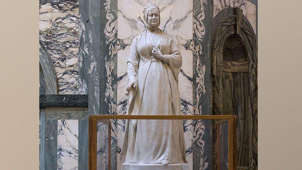 A statue in white marble of Elizabeth Fry. She is wearing a cap, a high-necked under shirt and a dress over the top caught up under the bust in the style of the first two decades of the 19th Century. The statue has a brass barrier around it and it is placed against a wall of swirling black and white marble