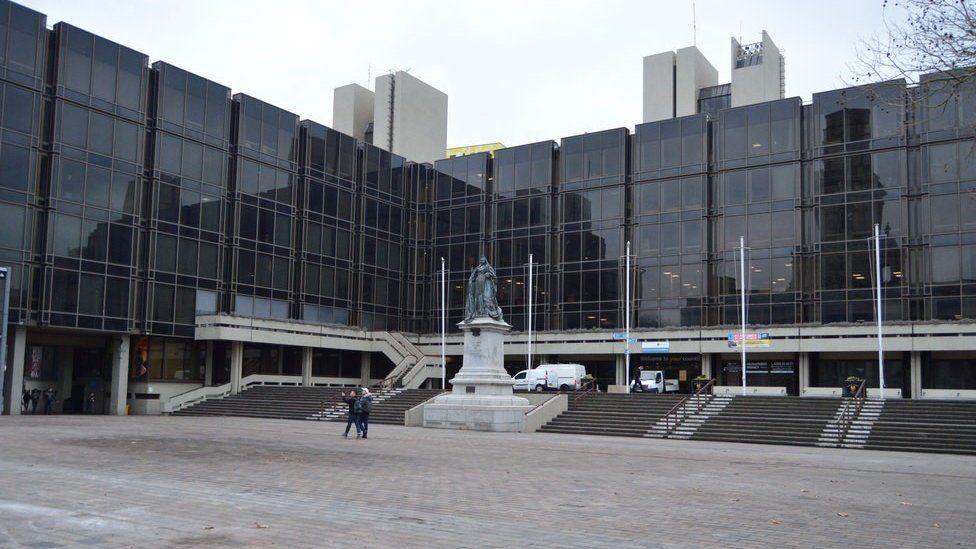 A large modern building with a glass frontage, with stairs leading up from a paved square and a statue of Queen Victoria on a plinth.