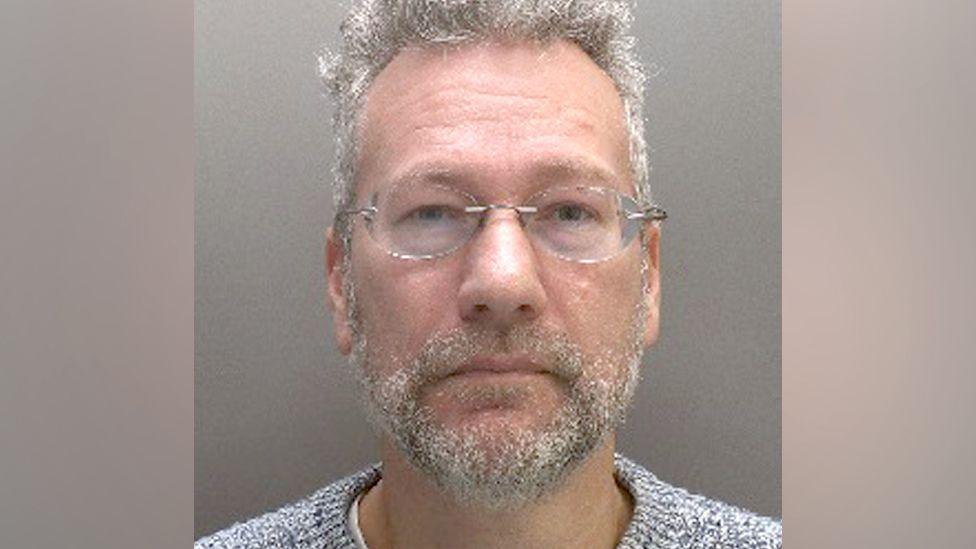 A mugshot of a man with grey hair and beard and glasses