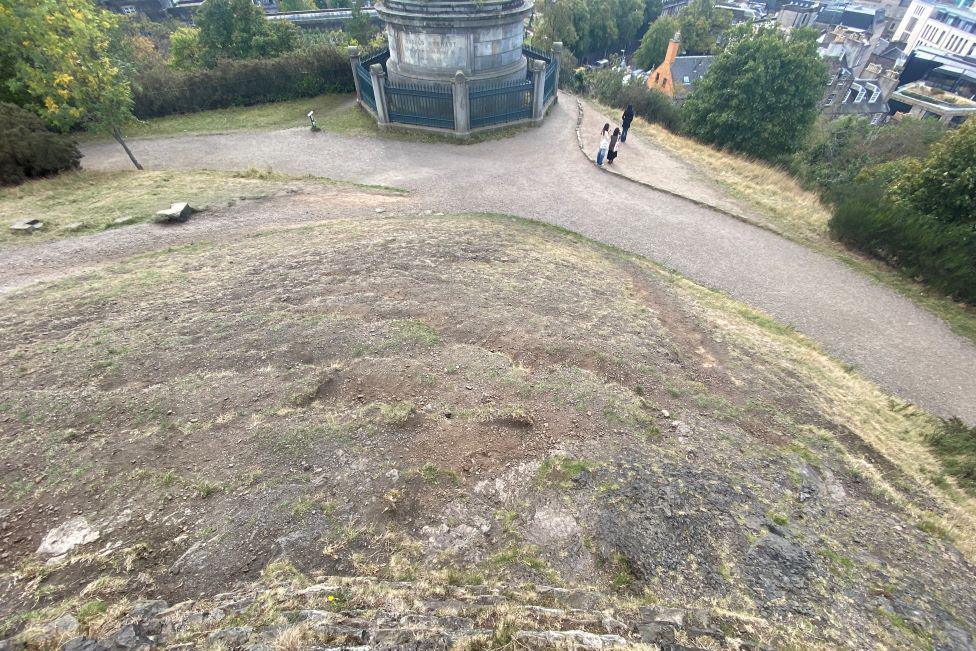 Looking down Calton Hill, a slope of bare earth is visible. There is evidence that the soil is slipping down the hill. Buildings in Edinburgh are visible in the background. 