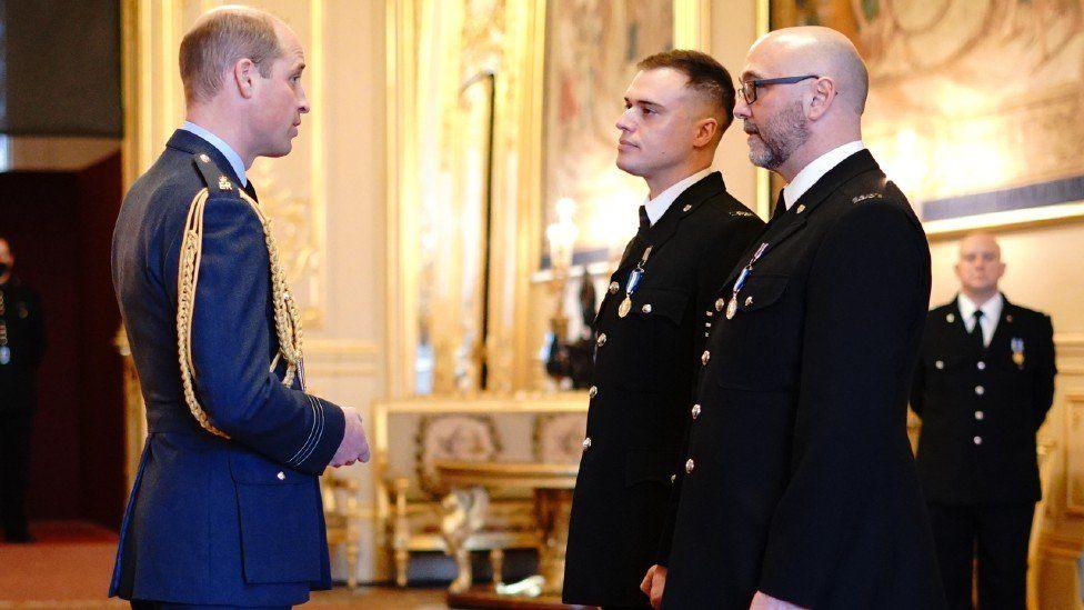 Sgt Michael Hooper and Constable Stephen Quartermain in their full uniform with navy jackets, white shirts and black ties, speaking to Prince William, wearing a navy uniform. Ornate white and gold furniture can be seen in the background.