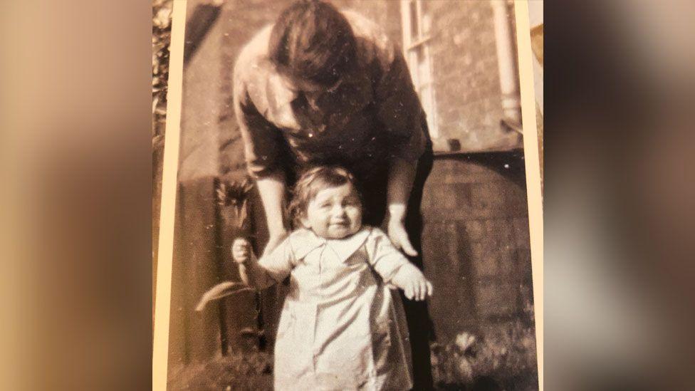 A black and white photo of Betty as a toddler holding a flower, she is being helped to stand by a woman who is behind her. 