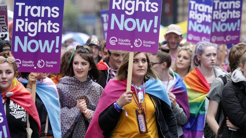 Pro-trans activists march with brightly coloured clothes, signs and flags 