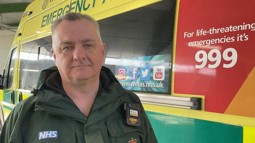 A male paramedic in uniform standing in front of an ambulance. Mr Raven is wearing a green ambulance uniform with a blue and white NHS badge on it.