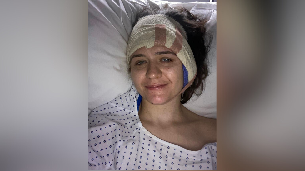 Suzi in a hospital bed. She is wearing a hospital gown with bandage around her head smiling at the camera.