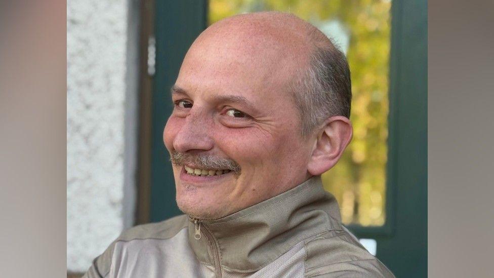 Arthur Polishchuk sits sideways-on to the camera and smiles. He is wearing a beige jacket that is zipped up. He has grey hair and a moustache.
