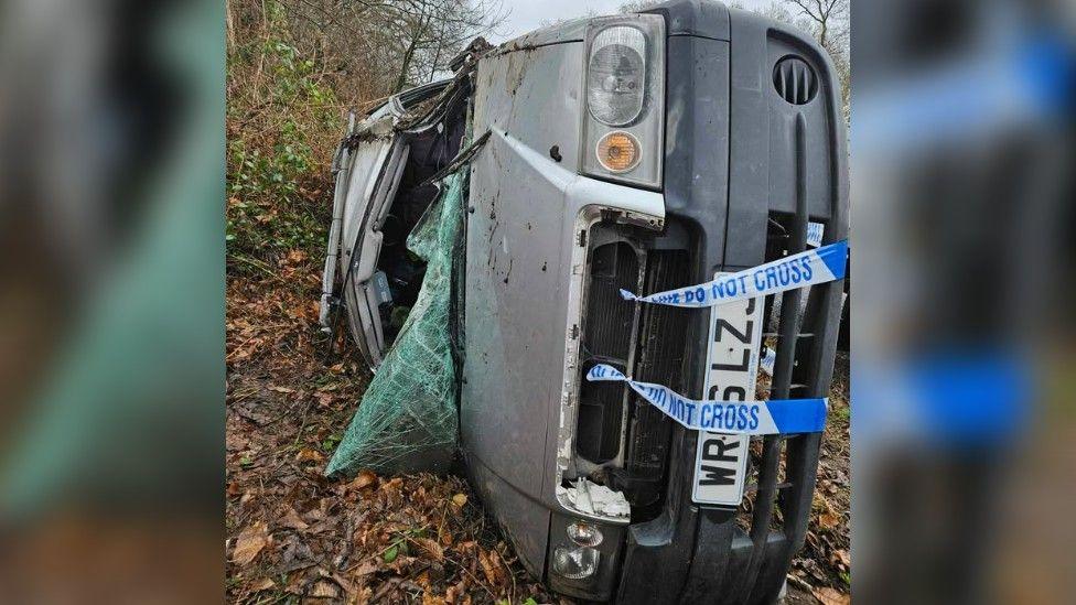 A damaged silver van lays on its side with a smashed, folded windscreen. It is among woodland. There is police tape wrapped around its bonnet.