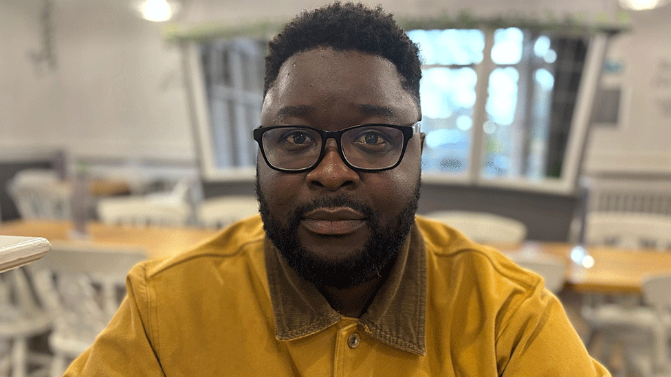 A man sits in a café wearing a mustard yellow shirt, looking at the camera. He has a beard and wears black-rimmed glasses