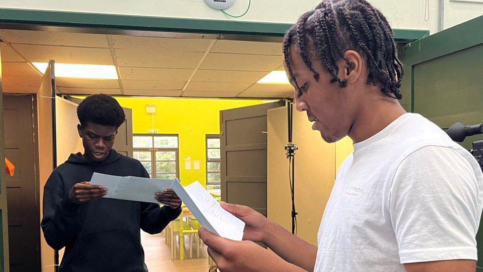 Two male students open their results and are looking down at their grades on pieces of paper in their school. The boy in the foreground is wearing a white T-shirt, while his friend in the background has a black jumper on.