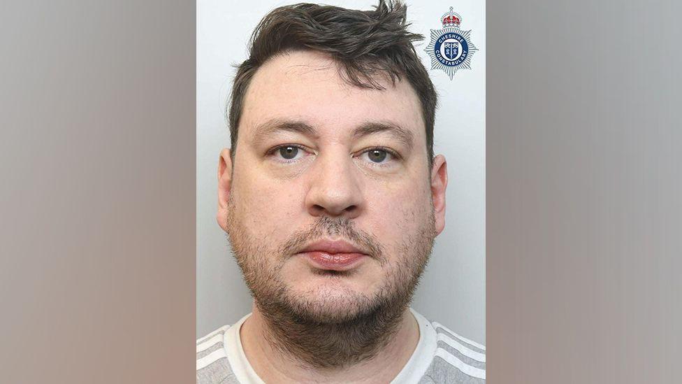 Thomas Hall faces the camera in a police mugshot. He has brown hair and facial stubble and is wearing a grey and white striped T-shirt.