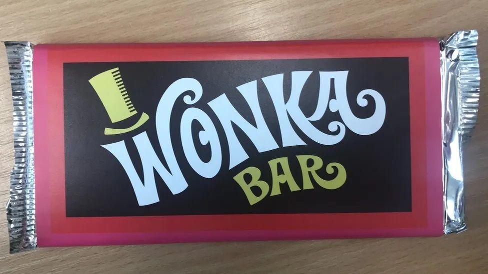 A chocolate bar labelled Wonka Bar with a top hat over the W and red and black packaging.