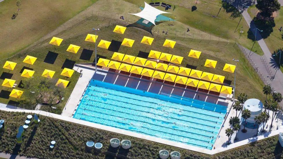 A computer-generated aerial image of an outdoor swimming pool with yellow sunshades that is surrounded by greenery, trees and pedestrian walkways.