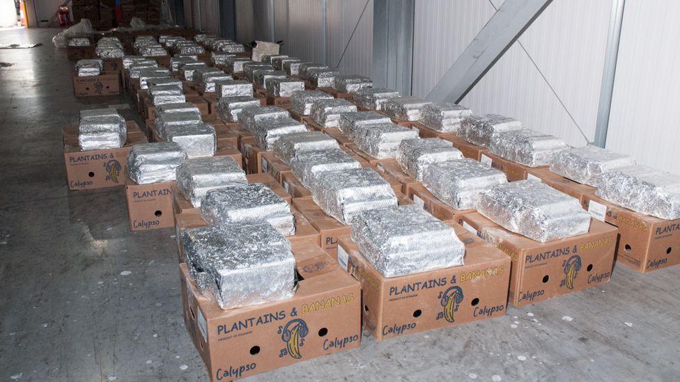 Dozens of cardboard boxes lined up on the floor of a warehouse. There are silver packages on top of every box.