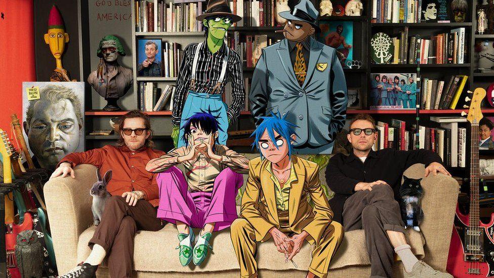 A picture of Gorillaz - including the animated bandmates - sitting on a sofa, surrounded by music instruments, figurines, celebrity photos, animals and a large bookcase at the back.