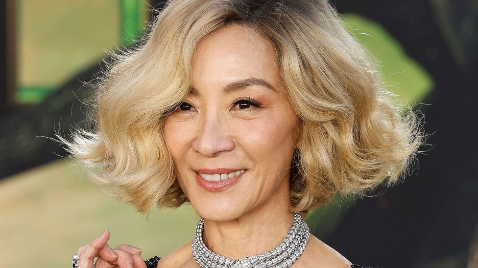 Michelle Yeoh with blonde hair and a thick, sparkling necklace, in front of a green Wicked-themed background at the film's premiere in Los Angeles