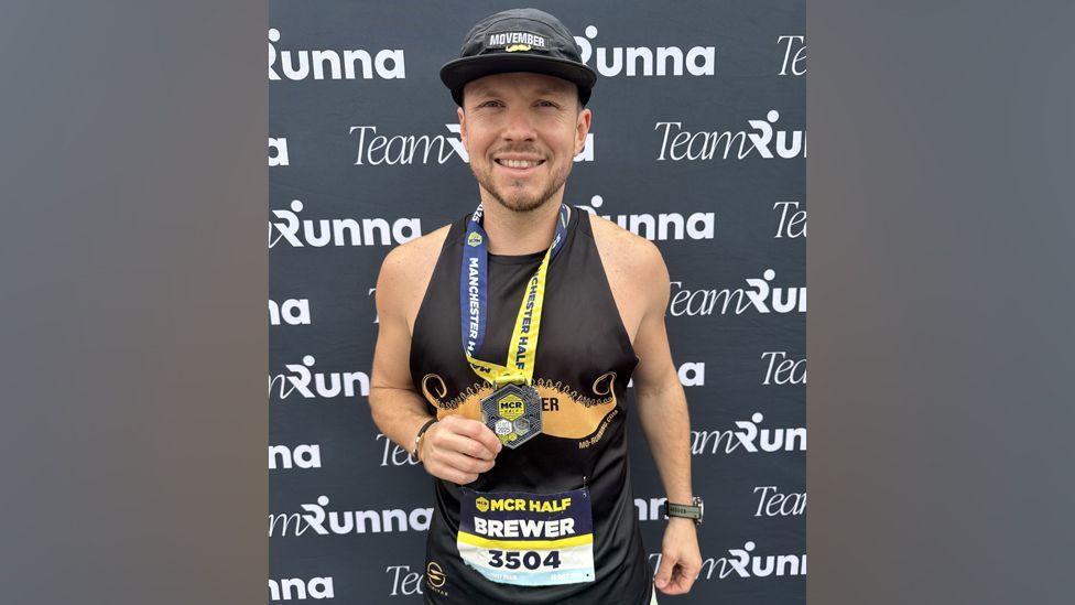 Bill Brewer, who has a running medal around his neck, is smiling at the camera and standing in front of an advertisement board. He is wearing a black cap and a black, sleeveless running top.