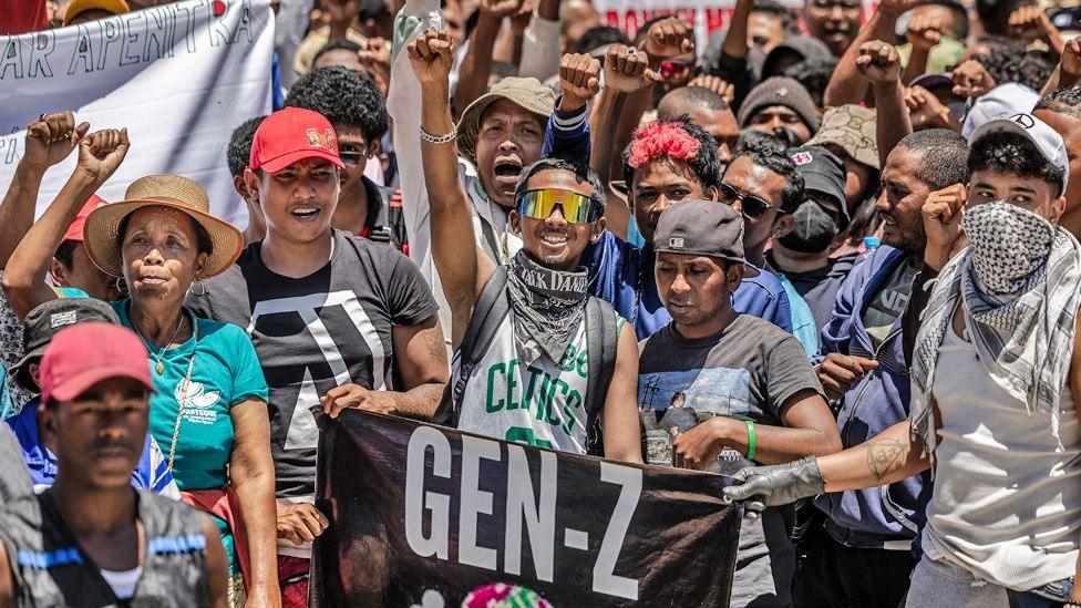 A group of protesters, some holding up a 'Gen Z' banner punch the air with their fists.