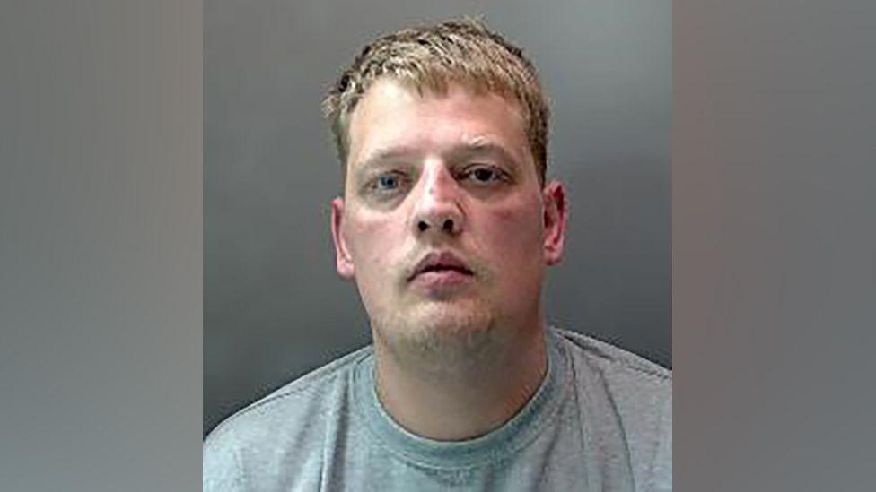A police custody photo of a man wearing a light grey crew-neck t-shirt against a plain grey background. He has short, neatly styled brown hair. The photo is taken from the shoulders up, with the person facing forward.