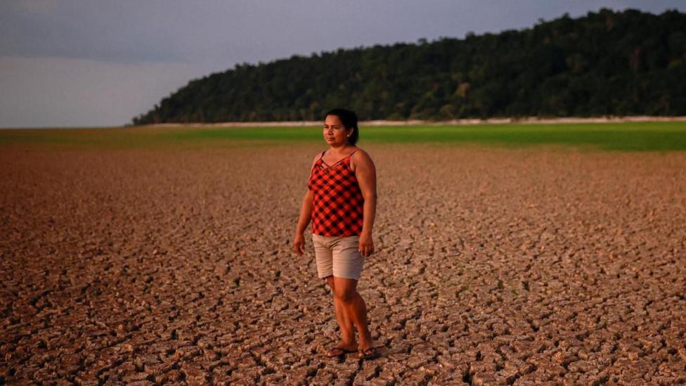 A woman in a red and black vest top, beige shorts and sandals stands on a dried-up river bed. There is a forest in the background.