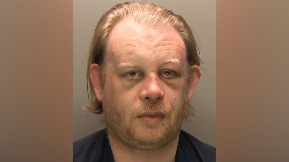 A mug shot of a man wearing a navy blue t-shirt. He has blond hair and a receding hairline. He also has blond facial hair.