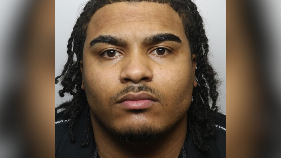 Zacharia looks at the camera with a blank expression. He is wearing a black top and has black braided hair, thick eyebrows and a thin moustache. 