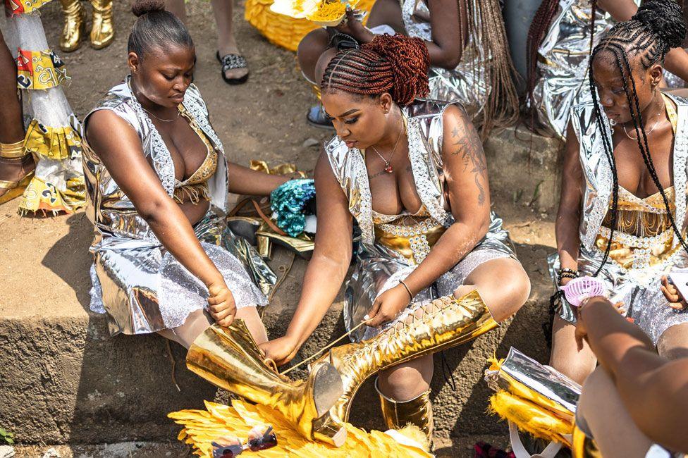 A woman in silver carnival skirt and waistcoat outfit helps a fellow participant in matching clothes lace up her gold boots - Calabar, Nigeria