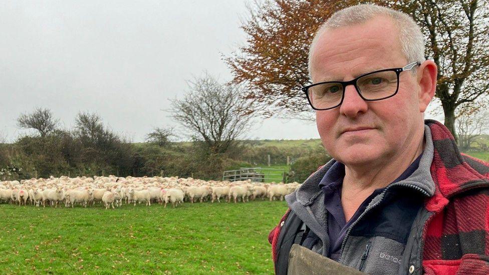 Farmer Colin Abel stands in the foreground wearing glasses, green overalls and a red and black checked overshirt. In the background a flock of sheep can be seen in a field.