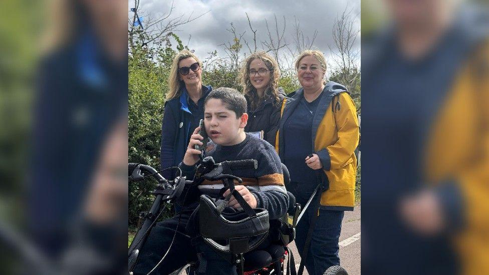 Jack on his trike, with a phone in his hand, with his mum, sister and family support worker from Julia's House