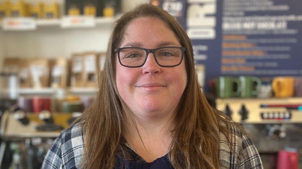 Grace Stewart a woman with long brunette hair. She is stood behind the counter at a cafe. She is wearing black rimmed glasses and is wearing a checked blue and white shirt with a blue apron.