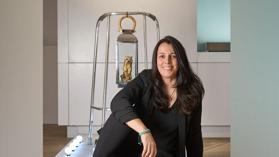 Maria Meyer sits in front of one of her exhibits - a Zimmer frame with a lantern hanging from the top, with a small sculpted dog inside it. Behind the exhibit is a cream wall with panels and a wooden floor.