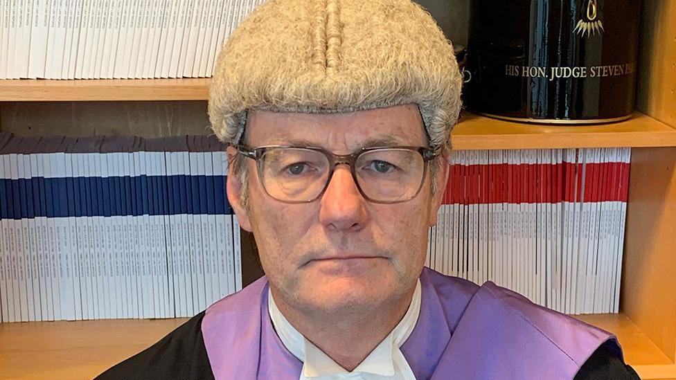 Judge Steven Parker, wearing wig and gown, looking at the camera in front of a bookshelf.