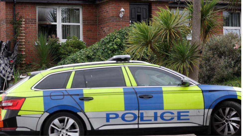 A marked police vehicle parked in front of a red brick house with damage to the front window