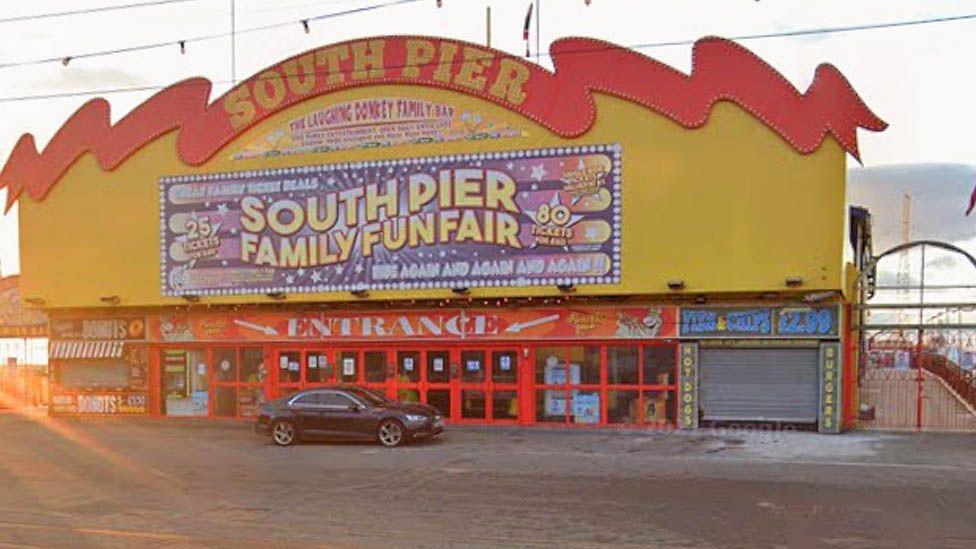 The entrance to South Pier, a large rectangular building painted yellow and red. It has a huge sign saying South Pier in yellow under a red roof shaped like a wave and many large red glass entrance doors with a small shop either side