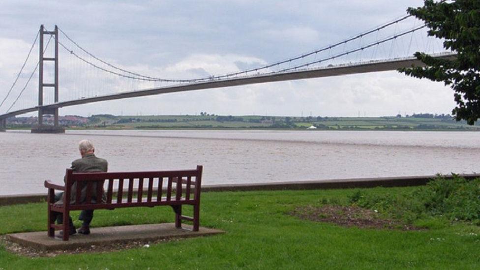 Humber Bridge viewing area on Hessle Foreshore, with a man sat on a bench on a grassy area next to the river 