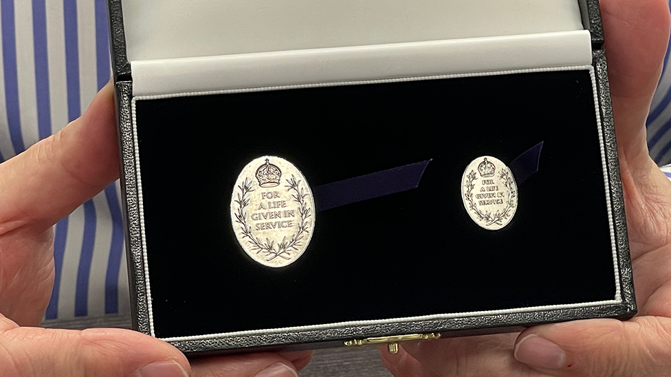 A silver-coloured, oval Elizabeth Emblem medal and accompanying miniature version being held in a black-lined case